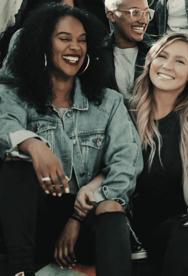 Alt tag: "Three friends laughing and enjoying a moment together, including a woman with curly hair, a person with short hair and glasses, and a woman with blonde hair, all casually dressed in a relaxed setting.