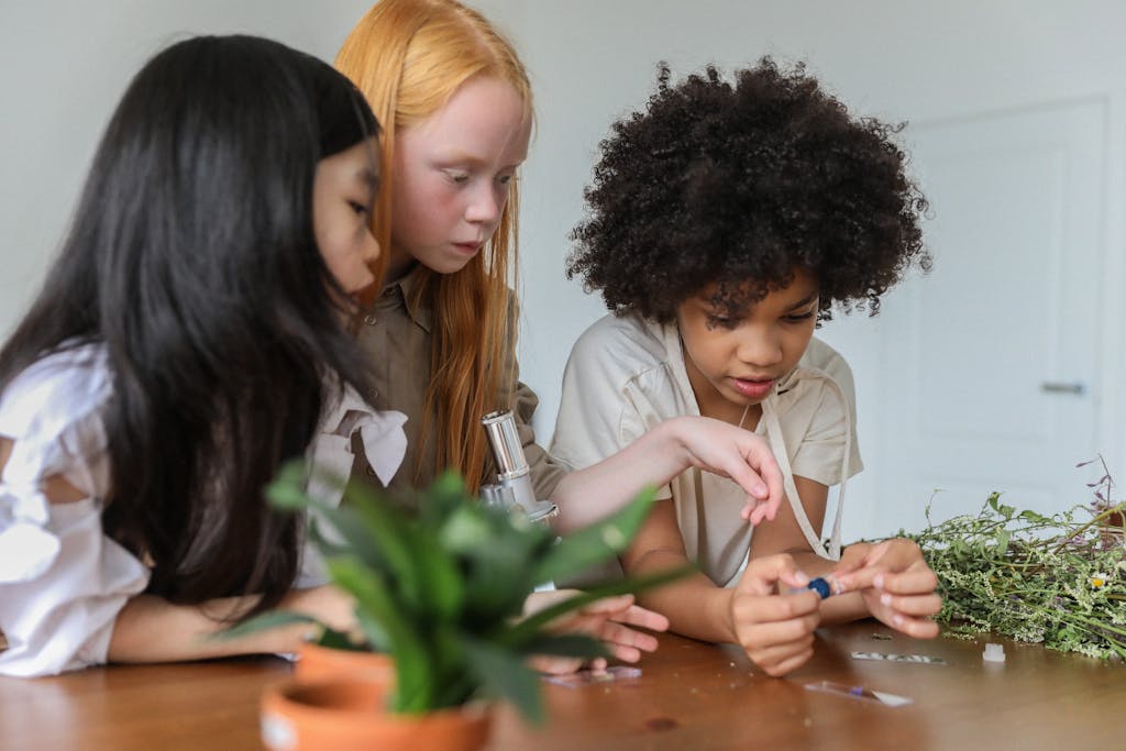 Concentrated multiracial children discussing while watching samples in microscope in room with plants