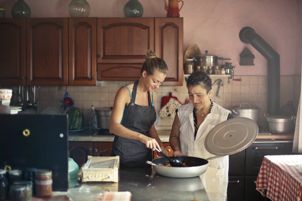 Daughter and senior mother standing at table in kitchen and stirring dish in frying pan while preparing food for dinner