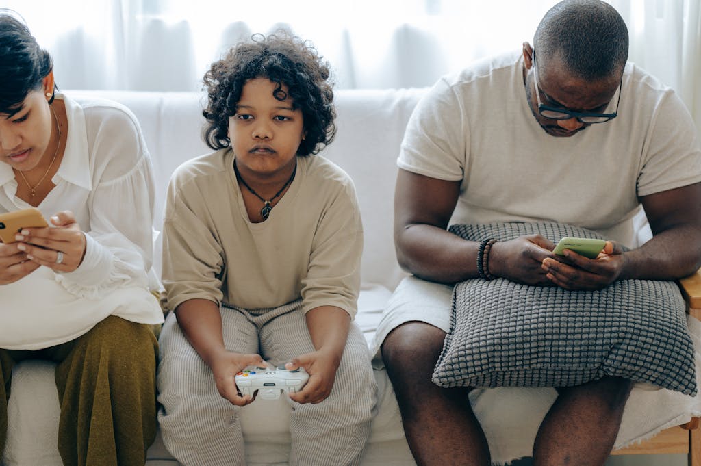 Focused ethnic child playing video game with joystick sitting on sofa between divers parents browsing smartphones
